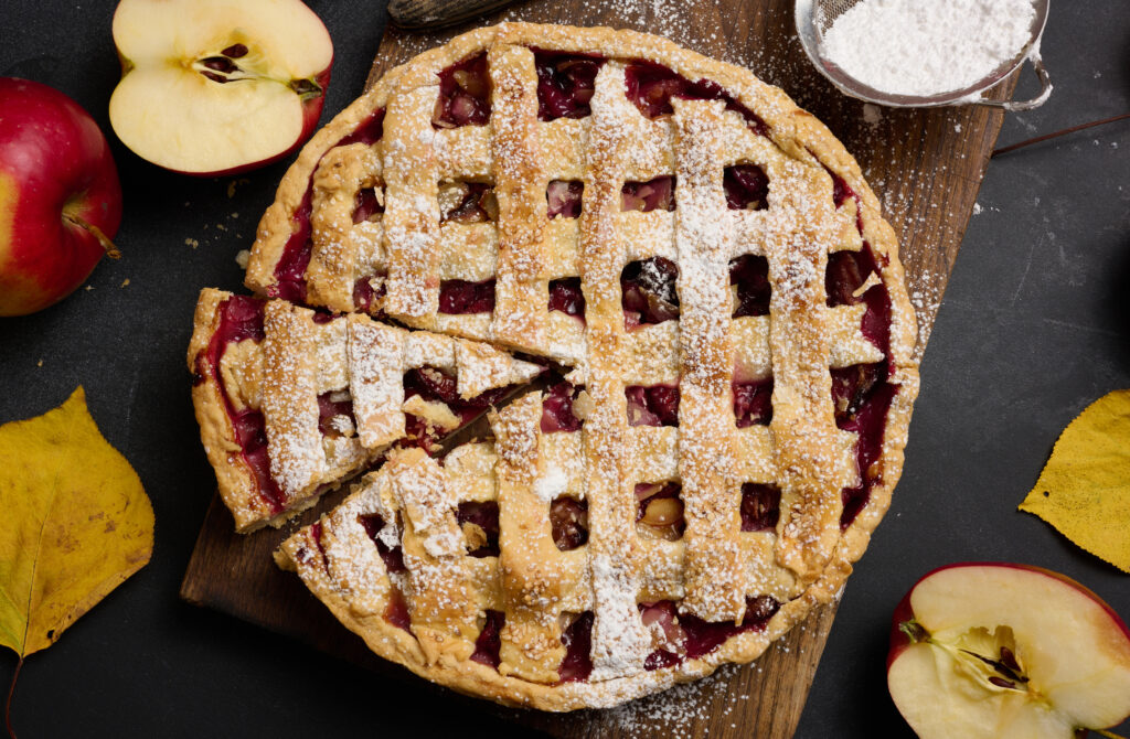 Baked round traditional apple pie on brown wooden board and fresh red apples, top view